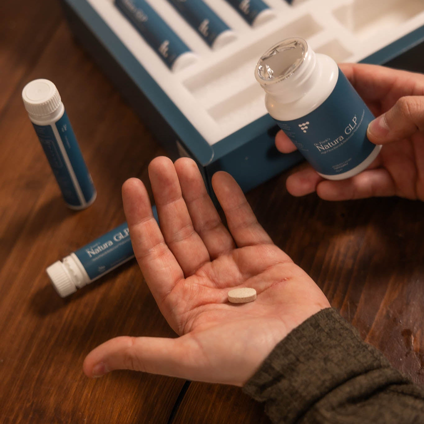 Person holding a supplement pill with a box of supplements in the background on a wooden surface.
